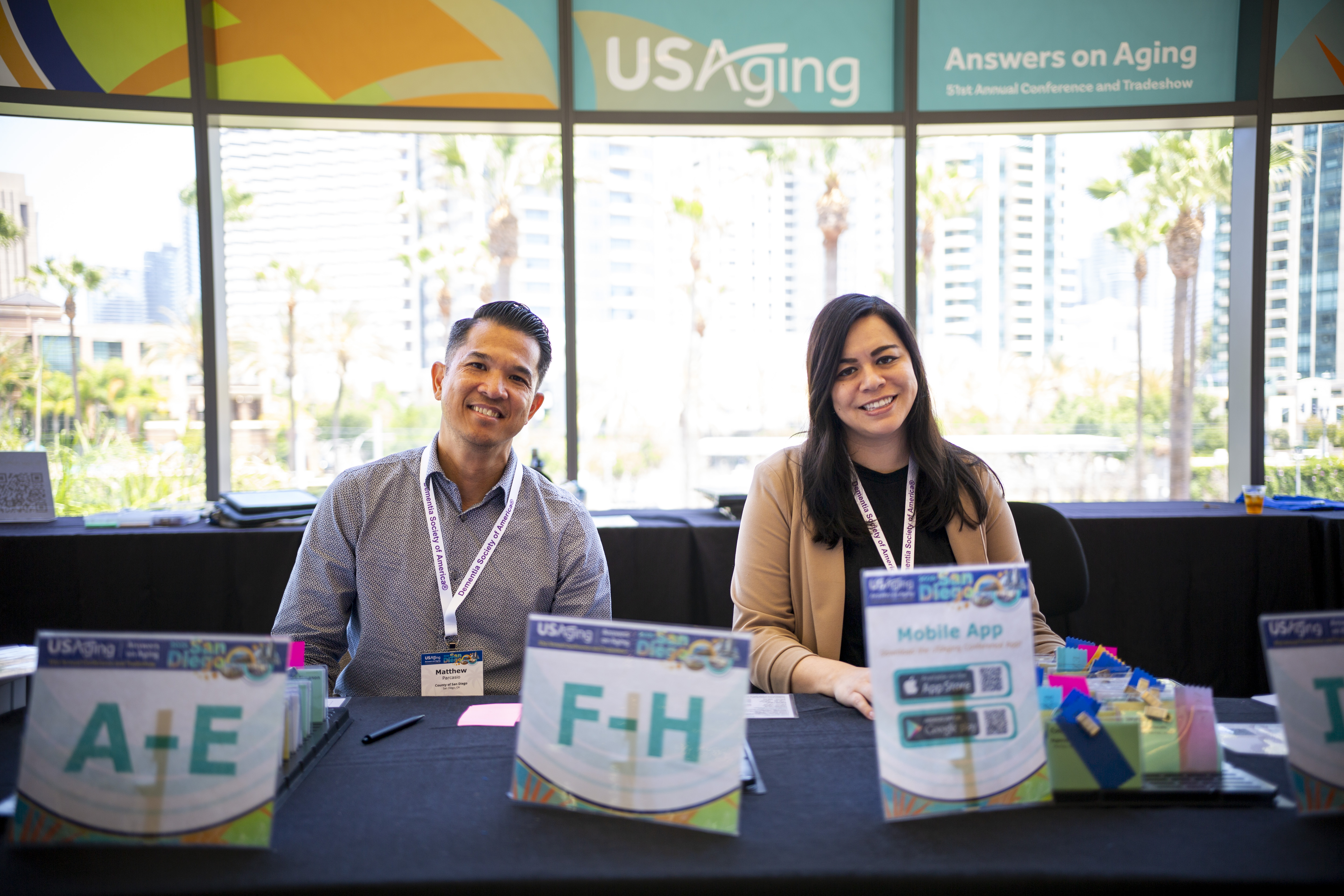 Two volunteers, one man and one woman, sit at the Registration table. The table has a black tablecloth, and there are small signs on the table with letters A to E and F to H on them, denoting where attendees should go to get their name badges. Large windows are behind them, with colorful USAging branded signs above the windows.